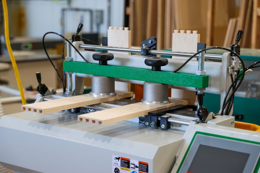 A woodworking machine precisely cuts dovetail joints into two light wooden boards, showing the careful craftsmanship we bring to each project in our welcoming workshop—guiding you every step of the way as your vision takes shape.